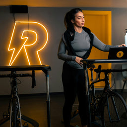 Woman with a backpack standing next to a bike in a room with a neon 'R' sign.