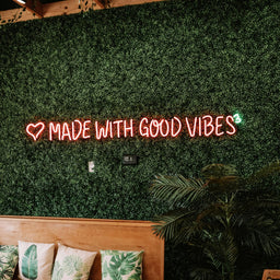 Neonkyltti 'MADE WITH GOOD VIBES' vihreällä seinällä, jossa on puinen penkki ja kasveja.
