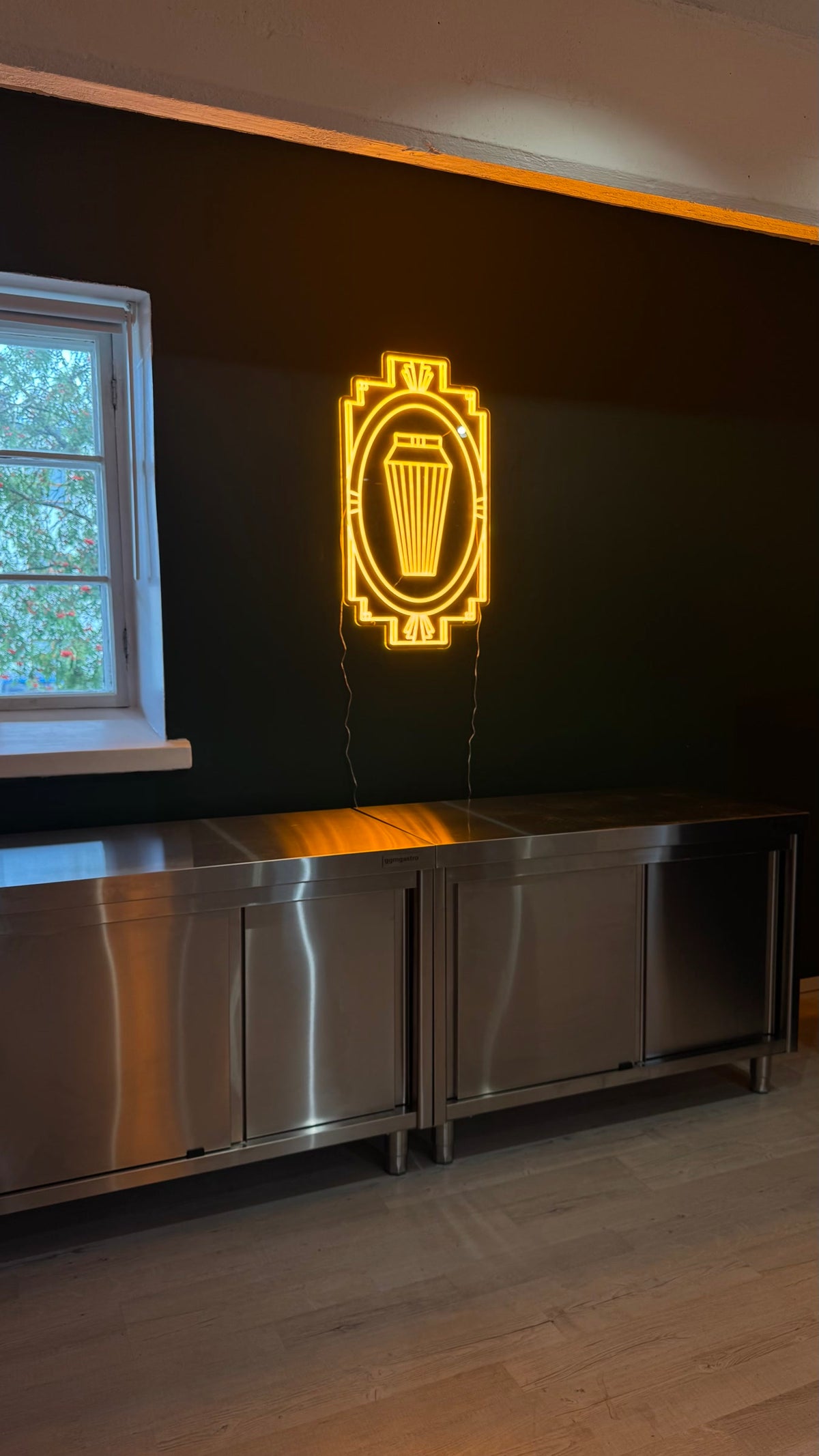 Lämpimänkeltainen cocktailshakerin muotoinen LED-neonvalo art deco -kehyksessä tummalla seinällä.