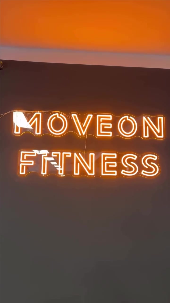 Lämpimän keltainen MOVEON FITNESS -LED-neonlogo tummalla liikuntastudion seinällä
